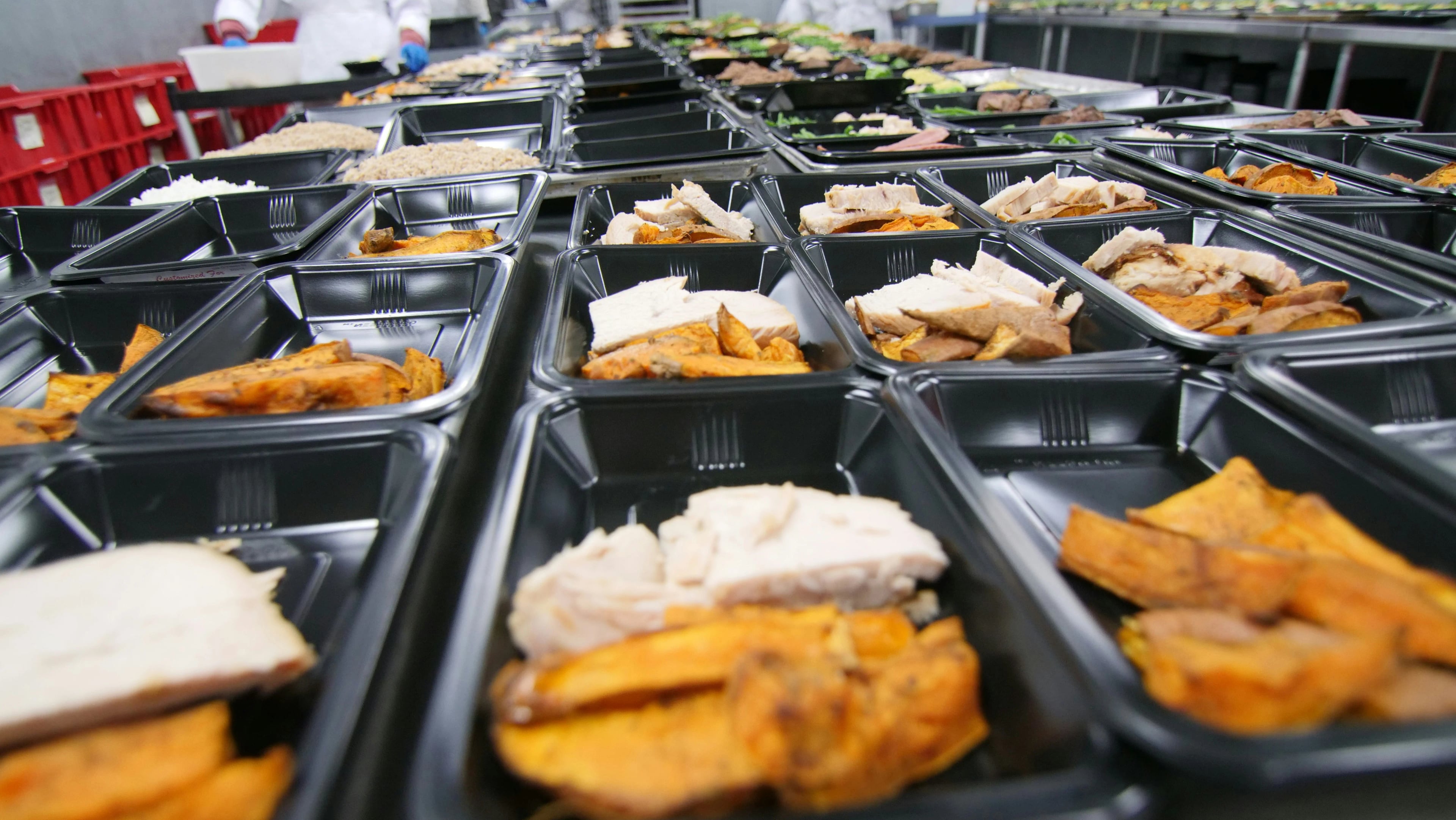 Rows of black meal prep containers filled with lean protein and roasted sweet potatoes on a commercial prep table.